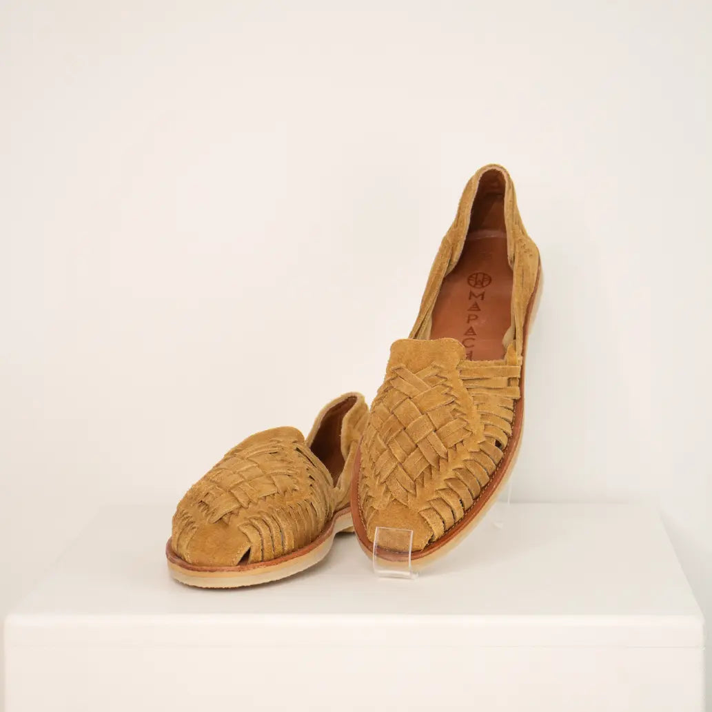Les Huaraches Rosalia Suede Camel sont posées de face sur une étagère blanche. La chaussure droite est relevée afin de mettre en valeur le tressage.