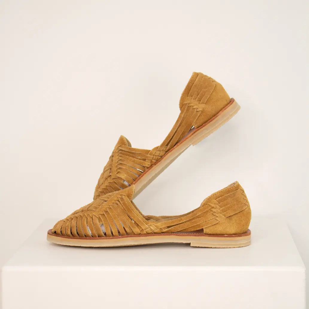 Les Huaraches Rosalia Suede Camel sont posées de profil sur une étagère blanche. La chaussure du fond est relevée afin de mettre en valeur le tressage.