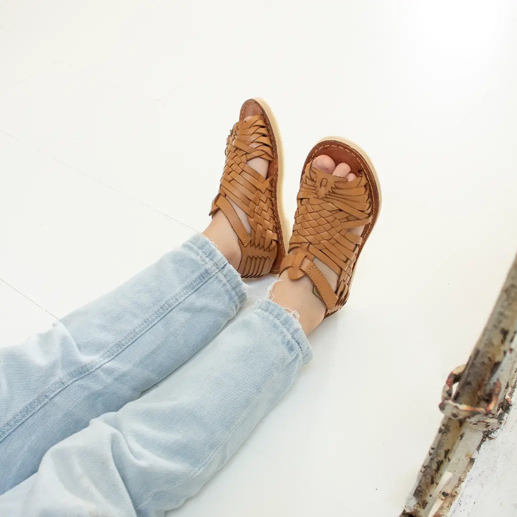 Une petite fille assise sur un sol blanc porte les Sandales Sandalita Camel ainsi qu'un jean bleu clair.