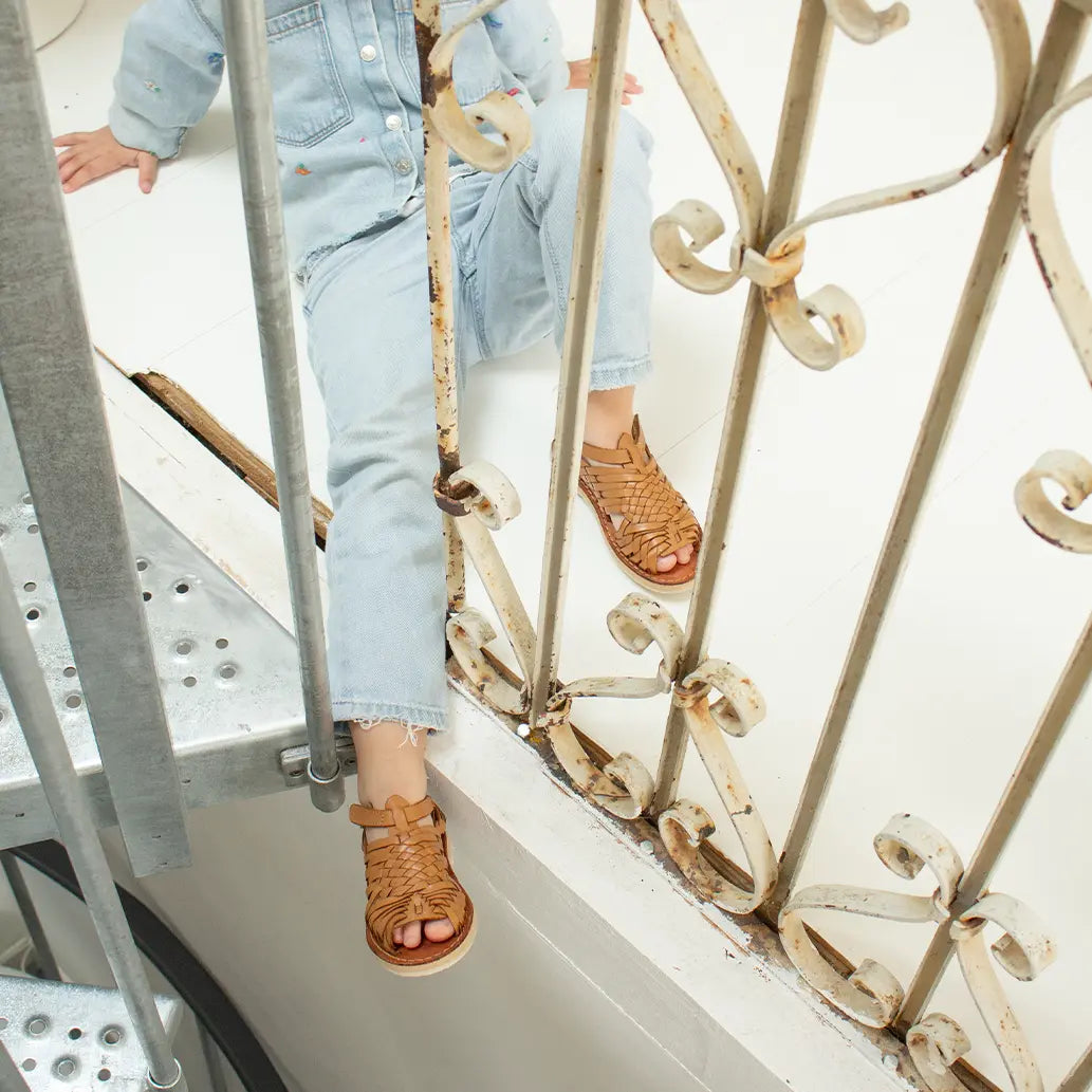 Une petite fille assise dans une cage d'escalier porte les Sandales Sandalita Camel ainsi qu'un ensemble pantalon chemise en jean.