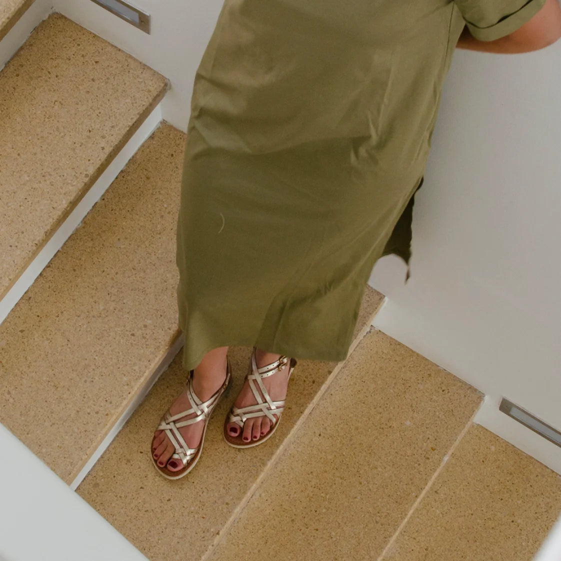 Une femme est debout dans des escalier en granit beige avec les Sandales Selena Champagne. Elle porte également une robe T-shit kaki.