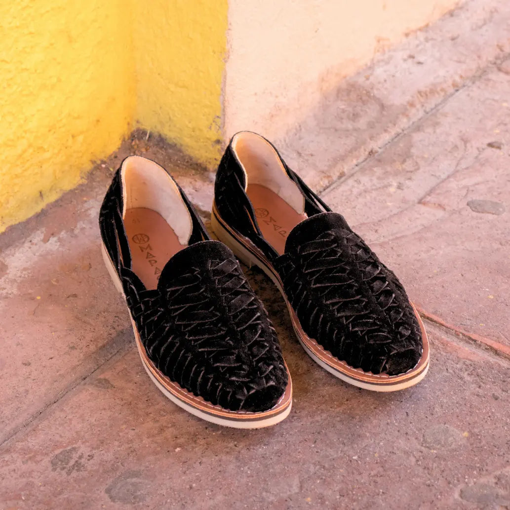 Les Mocassins Buena Vista Suede Noir sont posés sur un sol en terre rouge devant un mur jaune.