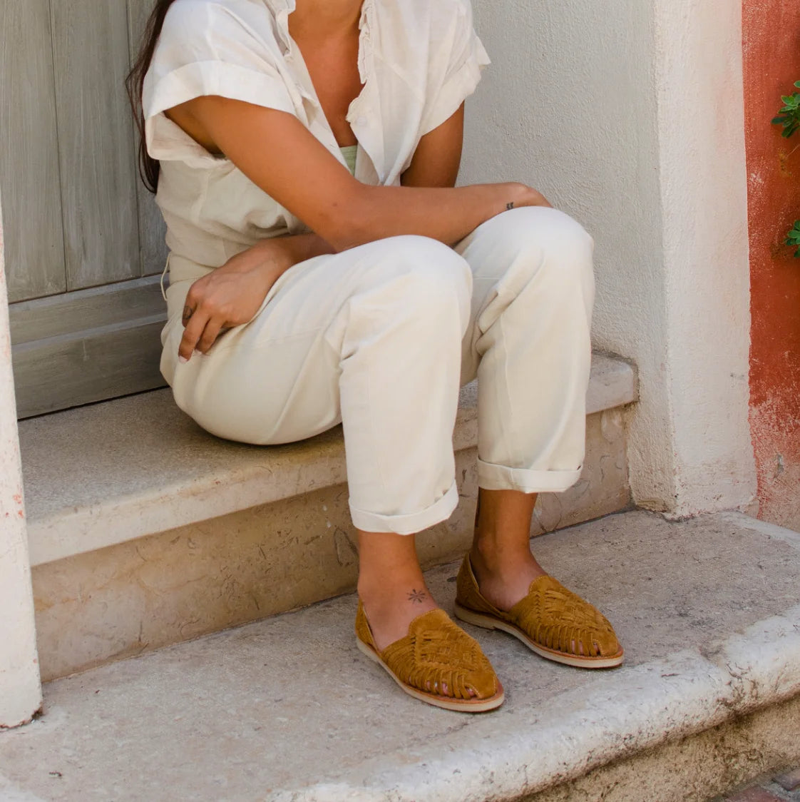 Une femme assise sur une marche devant une maison porte les Huaraches Suede Camel ainsi qu'une combinaison blanche.